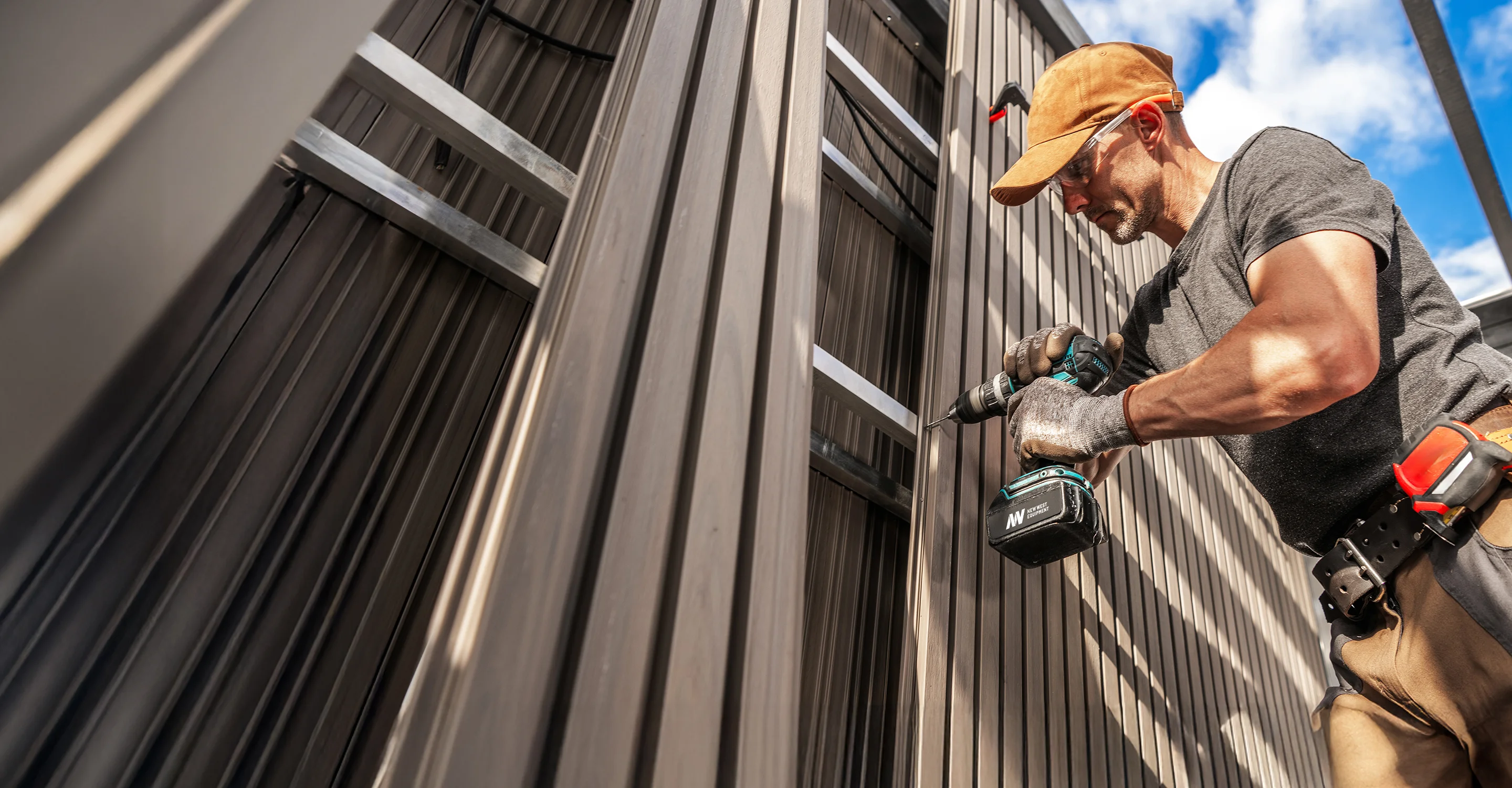 Construction worker in a cap and safety glasses using a cordless New West Equipment drill to fasten metal siding.