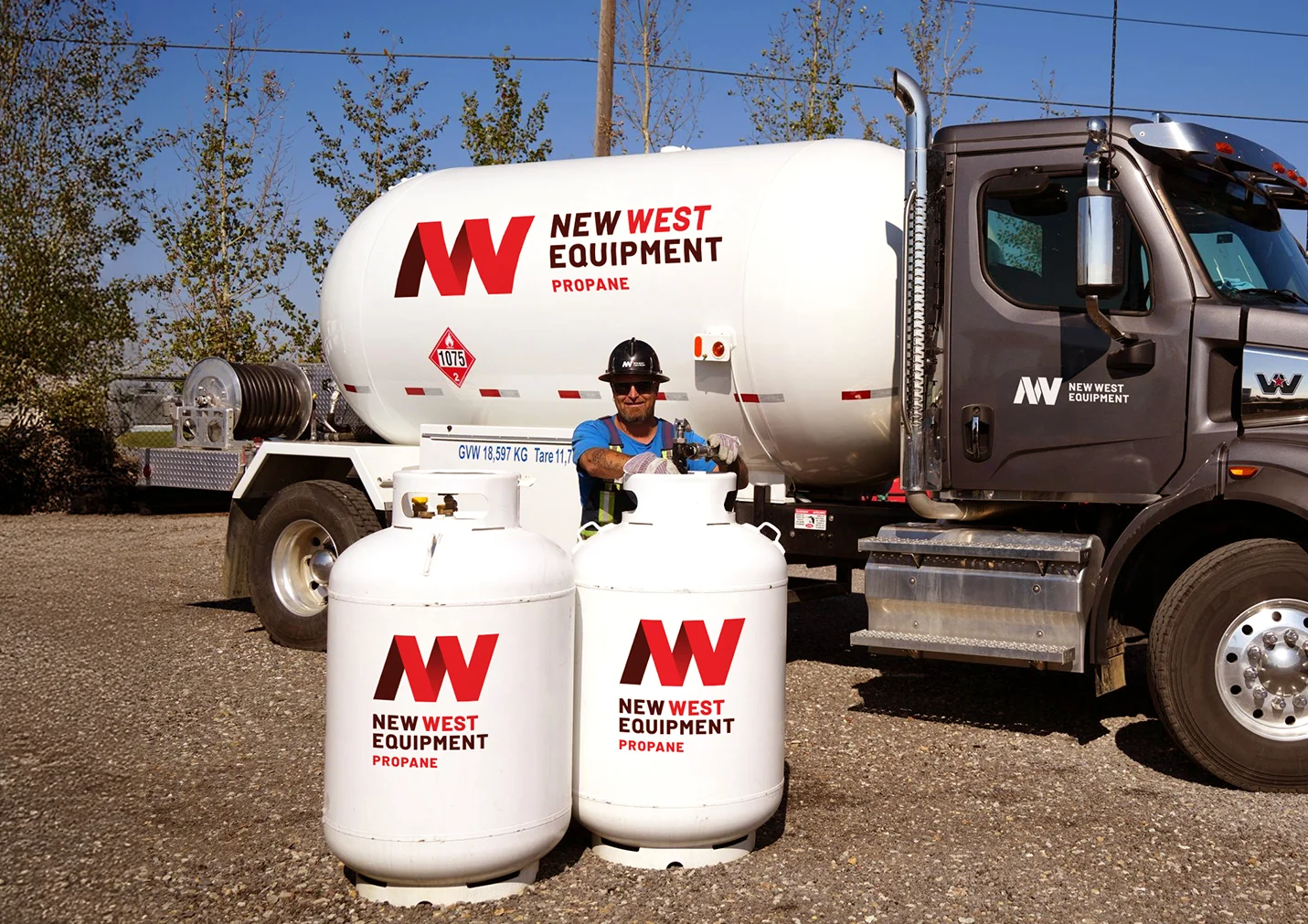 A worker in a hard hat and safety vest fills two white propane New West Equipment cylinders in front of a white tanker.