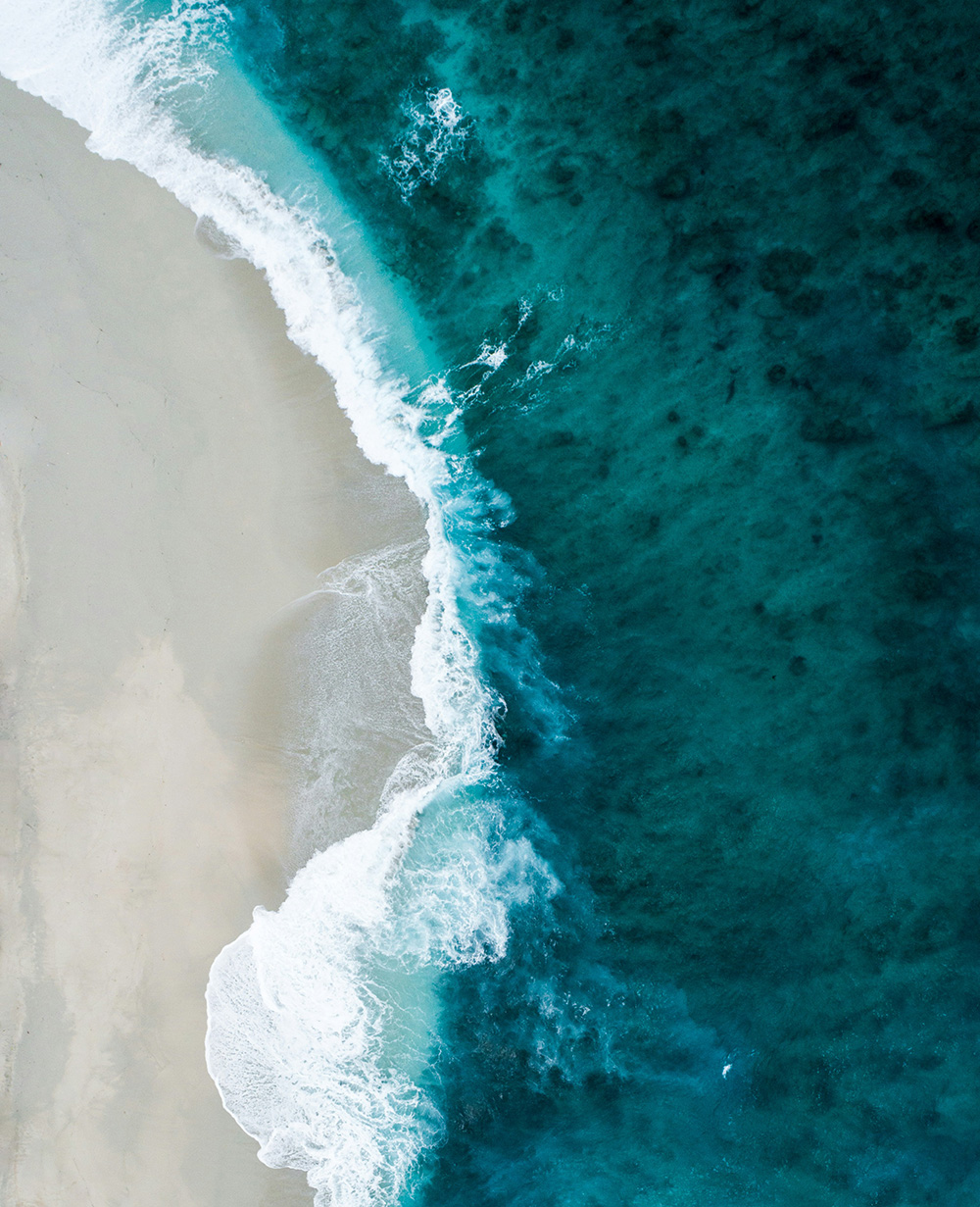 Aerial view of waves crashing onto a sandy beach with deep blue and turquoise ocean water.