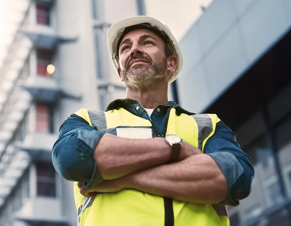 A confident construction worker wearing a white hard hat and yellow reflective vest stands with arms crossed in front of a modern building, looking upward.