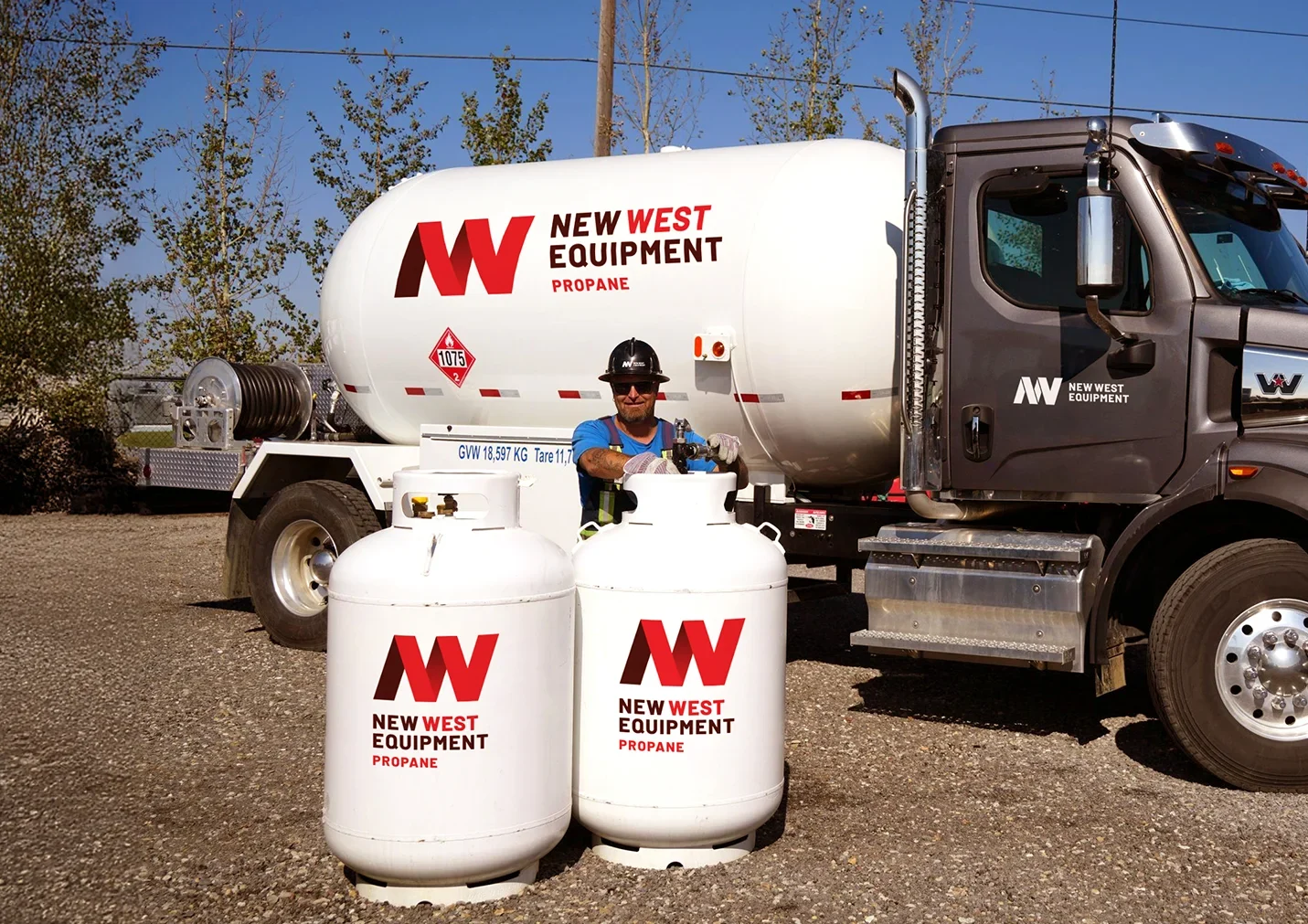 A worker in a hard hat and safety vest fills two white propane New West Equipment cylinders in front of a white tanker.