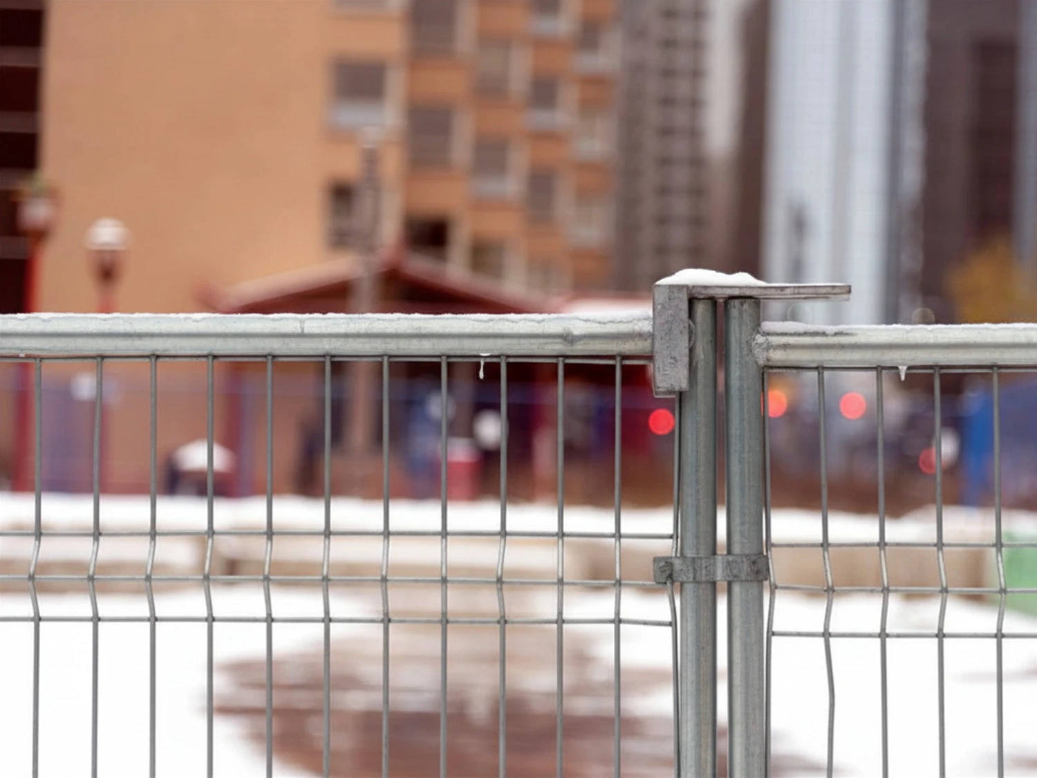 A close-up view of a metal fence with vertical and horizontal bars, secured with a bolt. In the blurred background, there are buildings and urban structures.