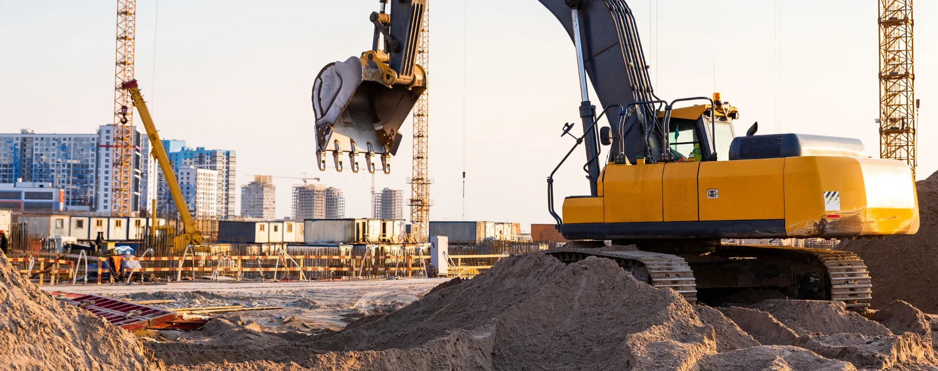 A yellow excavator is digging and moving soil at a construction site with partially built structures and cranes in the background. The scene captures active earthmoving work on the ground.